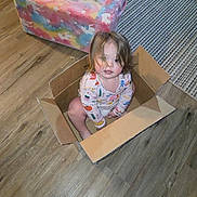 Jaylina joined the competition — help win amazing prizes! toddler, child, cardboard_box, floor, wooden_floor, plush_chair, colorful_chair, striped_rug, indoor, person, cute, curly_hair, looking_up, playful, home, casual_clothing, long_sleeve_shirt, barefoot, young_child, fun