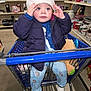 toddler, child, shopping_cart, santa_hat, plush_toy, jacket, leggings, socks, store, shelves, mugs, household_items, curious, indoor, winter_clothing, blue, pink, toy, retail, shopping