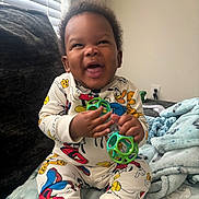 Rocket is registered to the contest to win money with this photo: baby, blanket, child, clothing, couch, cozy, curly_hair, cute, happy, indoor, infant, light, pajamas, person, playful, room, sitting, smiling, toy, window