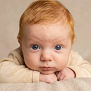 Maël a rejoint le concours — aidez-le/la à gagner de superbes lots ! baby, infant, blue_eyes, red_hair, closeup, portrait, face, hands, blanket, beige_background, soft_texture, cozy, neutral_expression, lying_on_stomach, fair_skin, onesie, eyelashes, forehead, studio_lighting, adorable