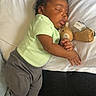 baby, sleeping, teddy_bear, bed, pillow, blanket, green_shirt, gray_pants, striped_socks, portrait, indoors, cozy, nap, cuddling, toy, bedroom, soft_lighting, dark_skin, infant, resting