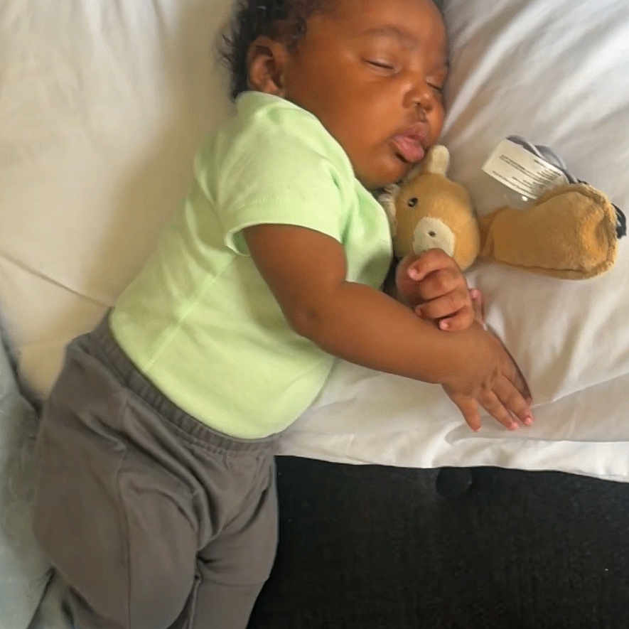Jazier joined the competition — help win amazing prizes! baby, sleeping, teddy_bear, bed, pillow, blanket, green_shirt, gray_pants, striped_socks, portrait, indoors, cozy, nap, cuddling, toy, bedroom, soft_lighting, dark_skin, infant, resting