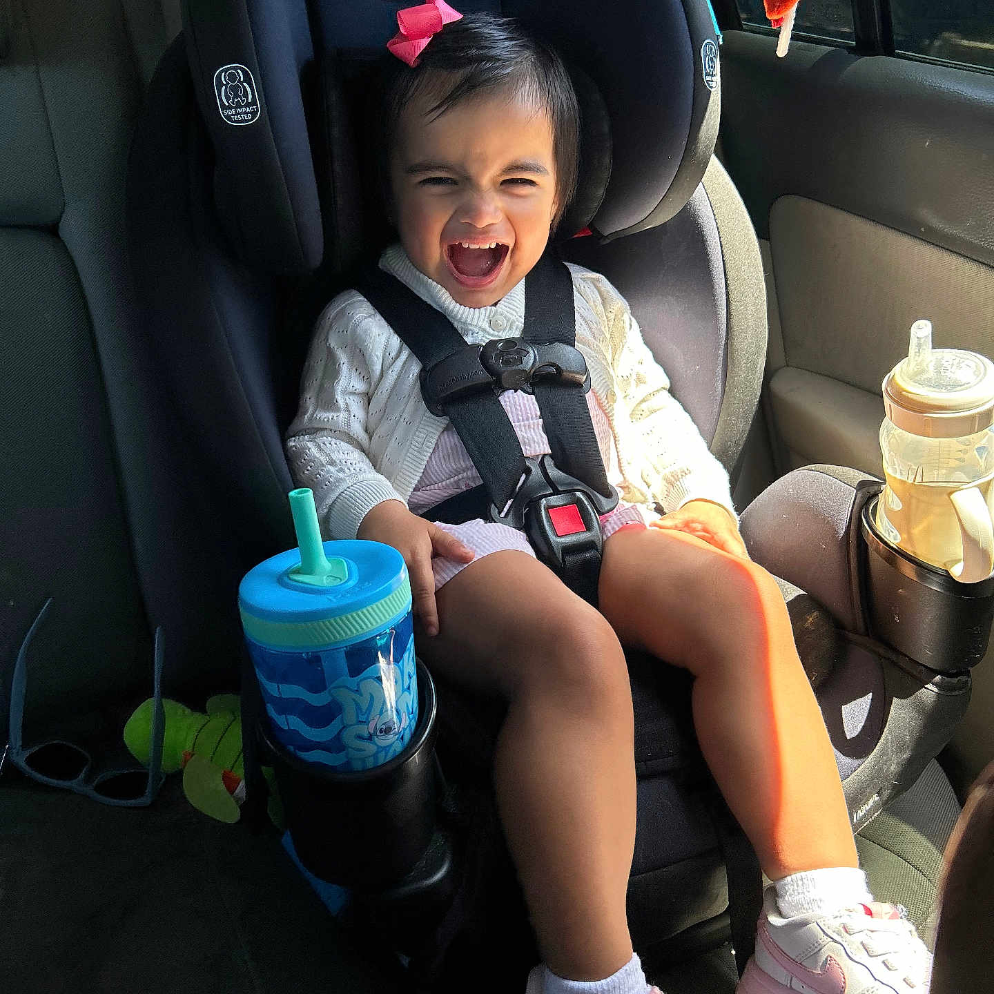 Avelinna is registered to the contest to win money with this photo: baby, car_interior, car_seat, car_trip, child, clothing, cup_holder, graco_brand, happy, pink_bow, seat, seatbelt, shoe, sippy_cup, smiling, socks, sunlight, toddler, toy, window