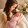 child, baby, newborn, girl, holding, swaddled, blanket, dress, long_hair, braid, flower_headband, indoor, portrait, family, love, cozy, soft_lighting, floral_background, warm_colors, sitting