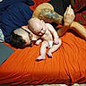 Daemon is registered to the contest to win money with this photo: baby, adult, tattoo, bed, orange_bed_sheet, diaper, glasses, indoor, person, lying_down, sitting, curious, casual, blanket, laptop, floor, socks, cozy, family, affection