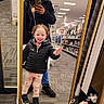 child, adult, mirror, store, aisle, jacket, pants, sneakers, smile, curly_hair, phone, reflection, carpet, shelf, product, lighting, casual, gesture, happy, shopping