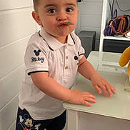 Juan participe au concours pour gagner de l'argent avec cette photo : bag, black_shoes, child, curious, expression, face, floor, hair, head, indoor, leaf, mickey_mouse, person, shorts, socks, standing, table, toddler, toy, white_shirt