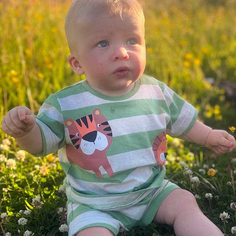 Tommy is registered to the contest to win money with this photo: baby, bodypart, clothing, face, finger, flower, grass, hand, head, herbal, herbs, person, photography, plant, portrait, shirt, shorts, sitting, sleeve, t_shirt