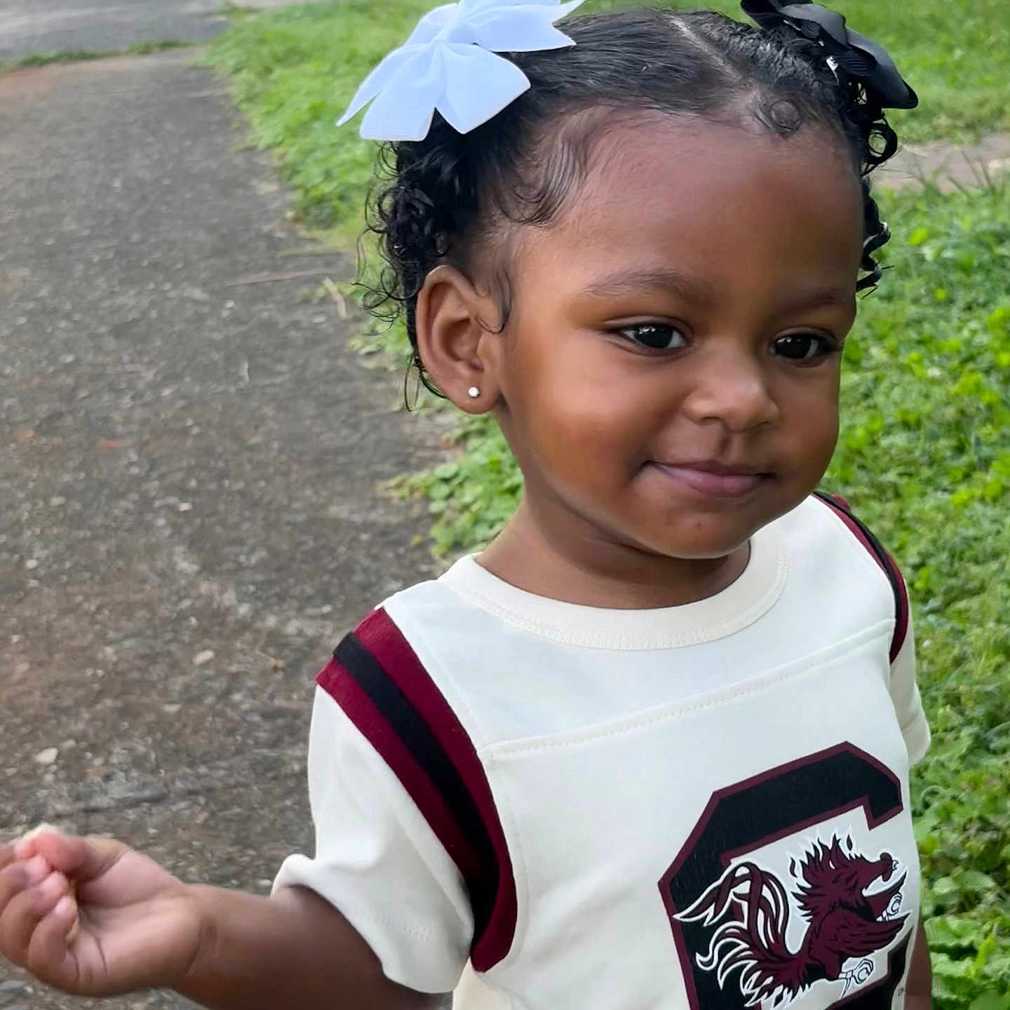 Khiari is registered to the contest to win money with this photo: car, casual_clothing, child, curly_hair, cute, daylight, earrings, emblem, grass, hair_bow, nature, outdoor, person, portrait, sidewalk, smile, toddler, walking, white_shirt, young_child