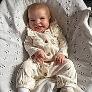 Isla is registered to the contest to win money with this photo: baby, smiling, onesie, knitted_blanket, shoes, white, cream, floral_pattern, happy, cute, infant, sitting, portrait, cozy, indoors, child, small_hands, soft_texture, clothing, fabric
