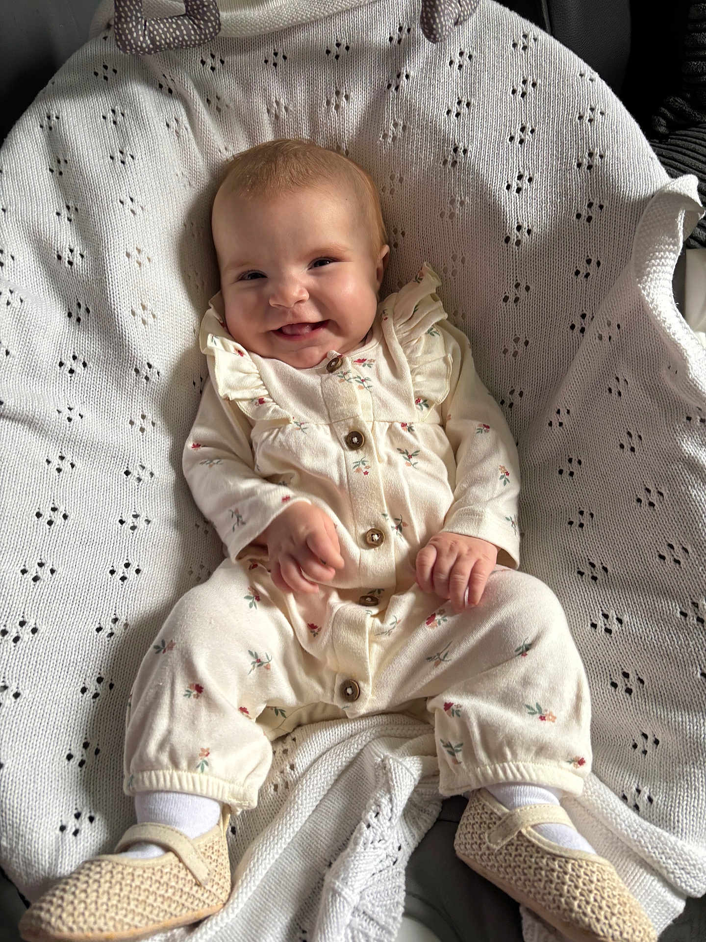 Isla is registered to the contest to win money with this photo: baby, smiling, onesie, knitted_blanket, shoes, white, cream, floral_pattern, happy, cute, infant, sitting, portrait, cozy, indoors, child, small_hands, soft_texture, clothing, fabric