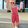 Stormy is registered to the contest to win money with this photo: child, toddler, pink_sunglasses, pink_romper, white_shoes, toy_case, outdoor, daylight, concrete, building, smiling, waving, shorts, curly_hair, casual_clothing, sunny, shadow, sidewalk, playful, happy