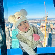 Abigail joined the competition — help win amazing prizes! bear_hat, blue_sky, child, cityscape, clothing, cute, fashion, fur_vest, happy, hat, indoor, mittens, playful, portrait, skyscraper, smiling, toddler, urban, window_view, winter_clothing