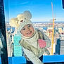 bear_hat, blue_sky, child, cityscape, clothing, cute, fashion, fur_vest, happy, hat, indoor, mittens, playful, portrait, skyscraper, smiling, toddler, urban, window_view, winter_clothing