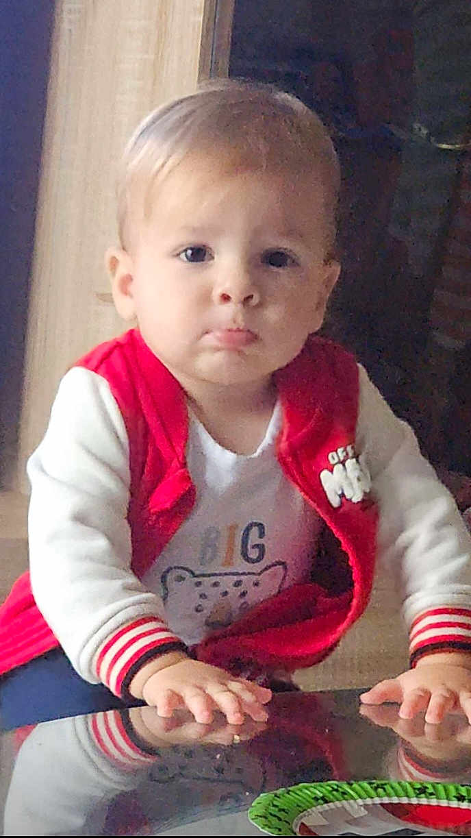 Nino participe au concours pour gagner de l'argent avec cette photo : toddler, child, face, red_jacket, white_shirt, cartoon_print, hands, glass_table, reflection, indoor, curious, pouty_expression, blond_hair, casual_clothing, young_child, person, portrait, sleeves, seated, home