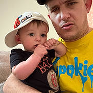 Lenny is registered to the contest to win money with this photo: baby, child, man, indoor, casual_clothing, cap, necklace, holding, curious, serious_expression, portrait, people, person, close-up, skin, arm, facial_expression, sitting, couch, family