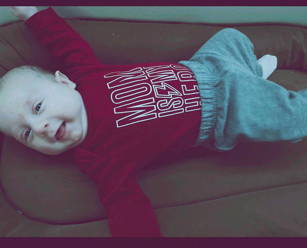 Elijah is registered to the contest to win money with this photo: baby, child, infant, smiling, red_shirt, grey_pants, couch, indoor, happy, face, person, cute, lying_down, casual_clothing, young_child, expression, portrait, comfortable, soft_surface, playful