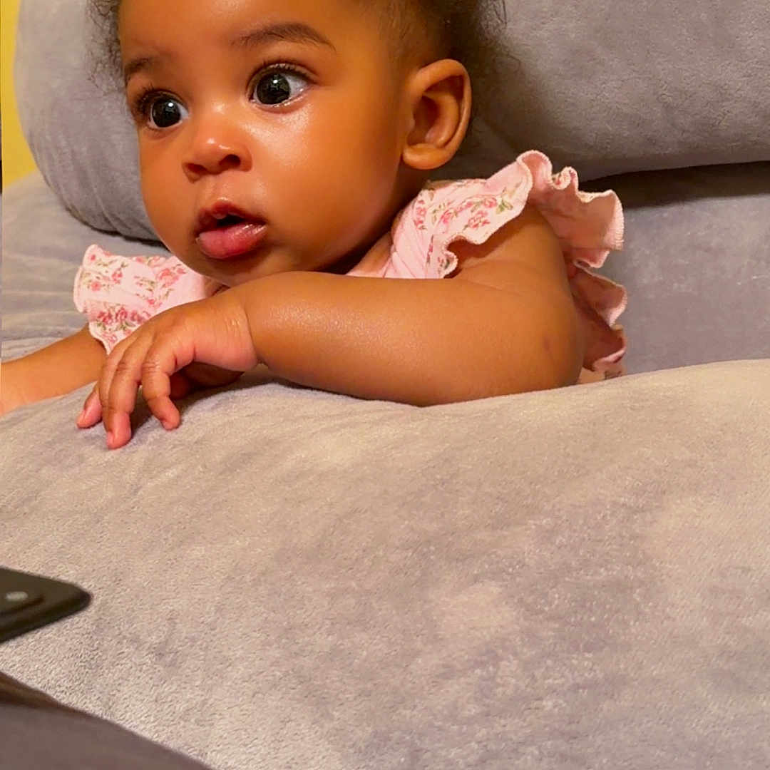 Emaan is registered to the contest to win money with this photo: baby, child, infant, curious, face, eyes, pink_clothing, floral_pattern, couch, soft_texture, yellow_wall, indoors, cute, portrait, skin, hair, hand, expression, seated, comfortable