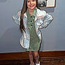 child, girl, smile, standing, long_hair, green_dress, plaid_jacket, white_shoes, wood_floor, blue_wall, framed_art, light_switch, indoor, casual_clothing, portrait, happy, hands_on_hips, home, fashion, young