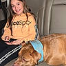 child, girl, dog, car_interior, seat, phone, hoodie, smile, pet, animal, leisure, casual_clothing, brown_dog, blue_collar, backseat, vehicle, young_person, happy, indoor, relaxed