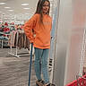 child, girl, hoodie, orange_clothing, ladder, shopping_cart, store, retail, clothing_rack, casual_pose, indoor, jeans, footwear, long_hair, smile, person, shopping, aisle, carpeted_floor, sneakers