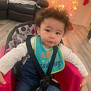 Osiah is registered to the contest to win money with this photo: toddler, child, curly_hair, bib, pink_chair, straps, christmas_tree, lights, living_room, leather_couch, stockings, flooring, denim_jeans, white_shirt, holiday_decor, baby_seat, indoor, person, cute, festive
