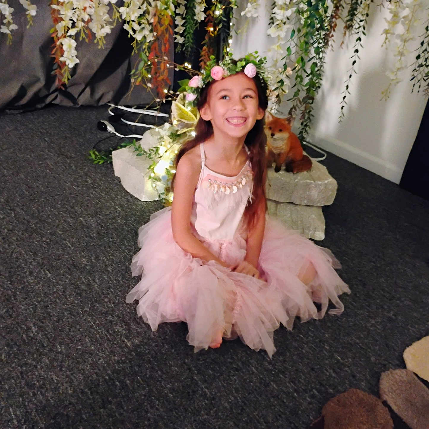 Omaris is registered to the contest to win money with this photo: carpet, child, costume, cute, decor, flower_crown, fox_figurine, girl, greenery, happy, indoor, lights, long_hair, pink_dress, playful, portrait, rocks, sitting, smiling, tutu