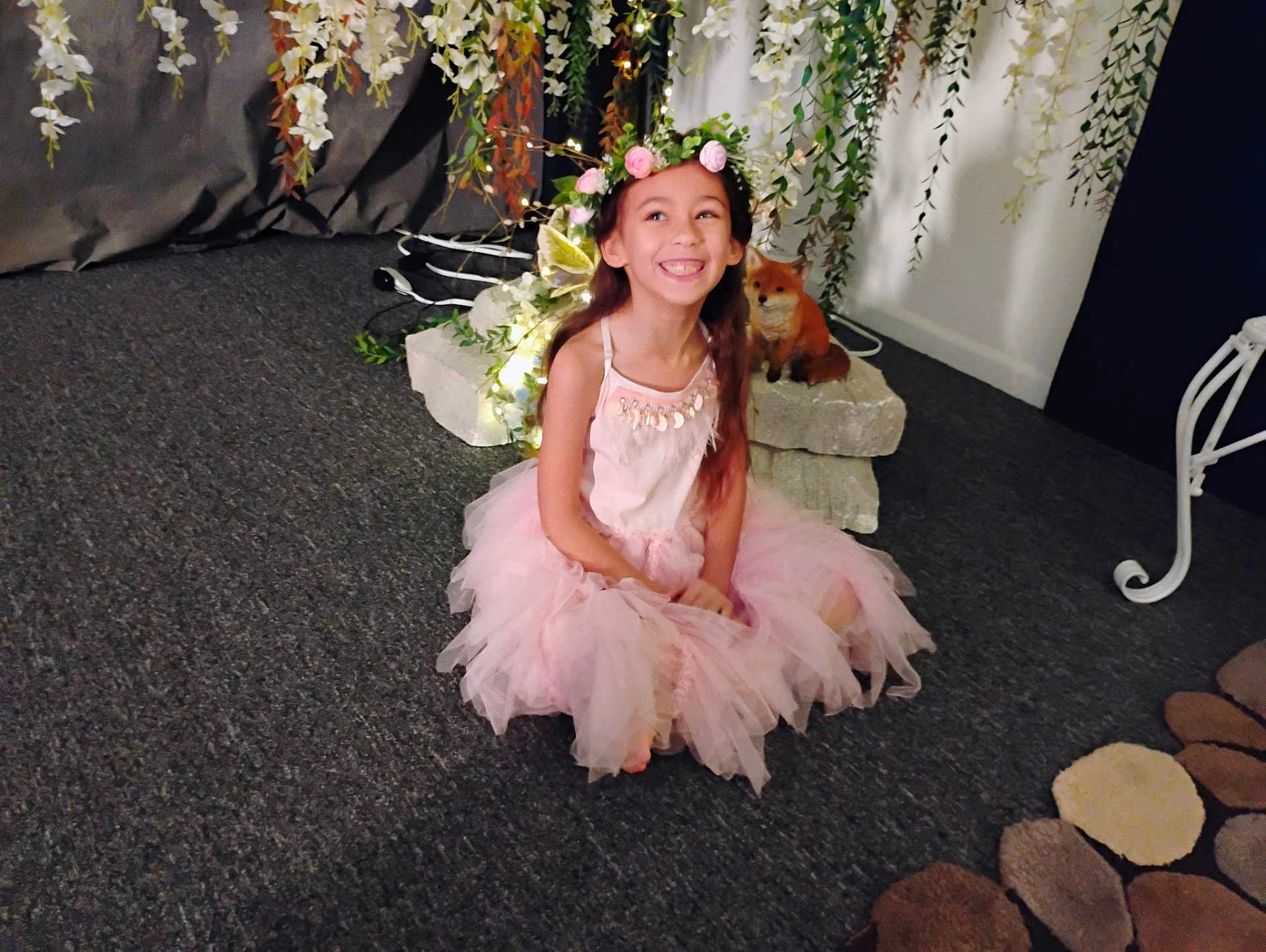Omaris is registered to the contest to win money with this photo: child, girl, smiling, tutu, pink_dress, flower_crown, indoor, carpet, fox_figurine, rocks, greenery, decor, lights, long_hair, happy, sitting, playful, costume, portrait, cute