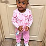 beige_door, braided_hair, casual_wear, child, curious_expression, cute, door, earrings, floor_tiles, hair_bow, hello_kitty, holding_object, indoor, pink_clothing, portrait, sneakers, standing, toddler, water_bottle, young_child