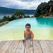 Souleyman a rejoint le concours — aidez-le/la à gagner de superbes lots ! child, toddler, pool, water, wooden_deck, swimming_trunks, outdoor, nature, lake, trees, cloudy_sky, summer, curly_hair, person, young_child, water_play, recreation, vacation, daylight, relaxation