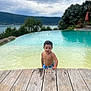 child, toddler, pool, water, wooden_deck, swimming_trunks, outdoor, nature, lake, trees, cloudy_sky, summer, curly_hair, person, young_child, water_play, recreation, vacation, daylight, relaxation