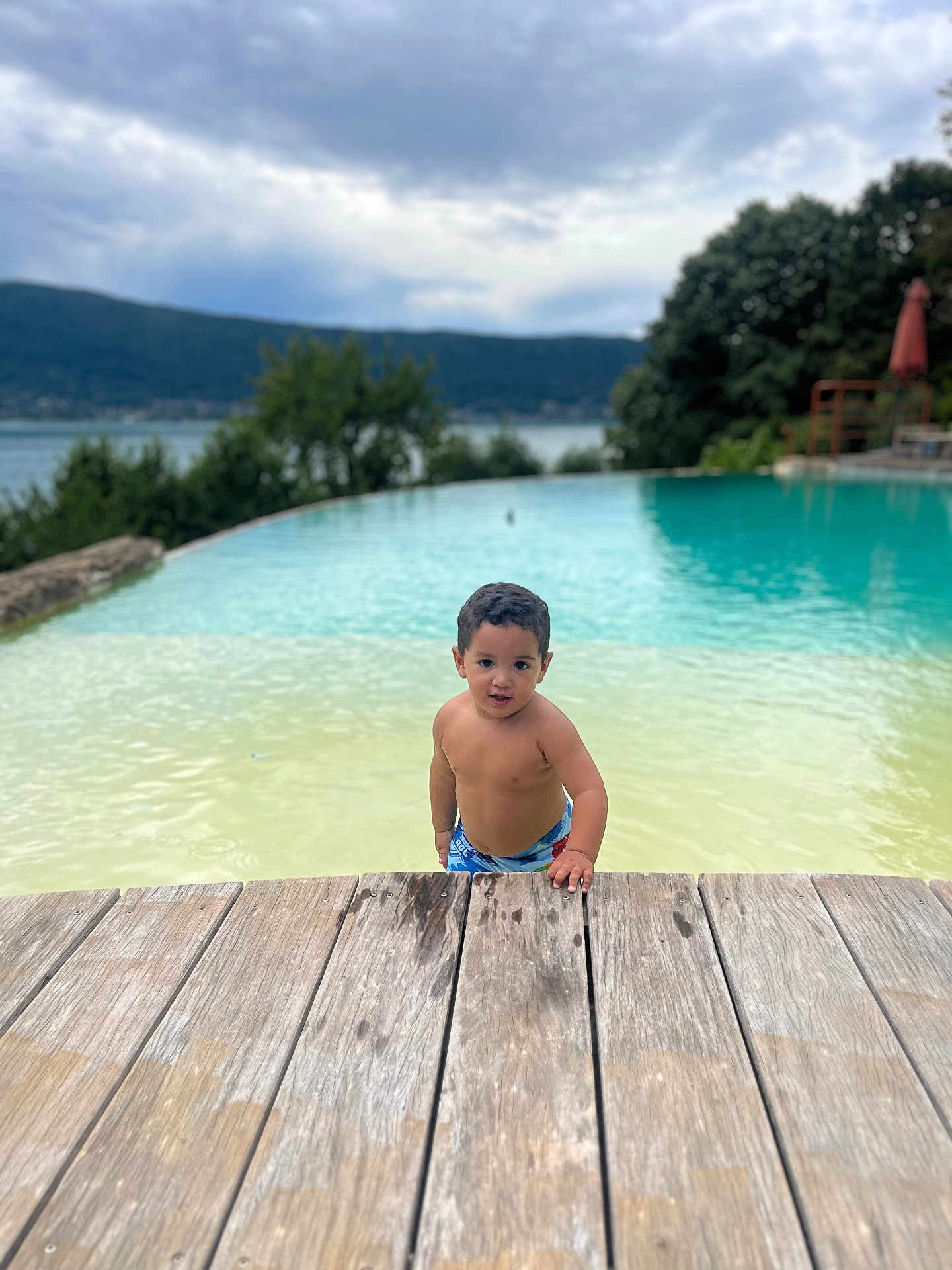 Souleyman a rejoint le concours — aidez-le/la à gagner de superbes lots ! child, toddler, pool, water, wooden_deck, swimming_trunks, outdoor, nature, lake, trees, cloudy_sky, summer, curly_hair, person, young_child, water_play, recreation, vacation, daylight, relaxation