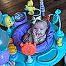 baby, child, infant, smiling, happy, tongue_out, bow_headband, activity_center, baby_toy, colorful, playtime, indoor, wood_floor, seated, portrait, face, toy_robot, plush_toy, plastic_toys, family_photo