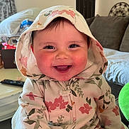 Evelyn joined the competition — help win amazing prizes! baby, child, smiling, hoodie, floral_pattern, rosy_cheeks, indoors, living_room, couch, plush_toy, table, remote_control, happy, face, person, cute, cozy, hood, expression, portrait