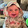 baby, child, smiling, hoodie, floral_pattern, rosy_cheeks, indoors, living_room, couch, plush_toy, table, remote_control, happy, face, person, cute, cozy, hood, expression, portrait