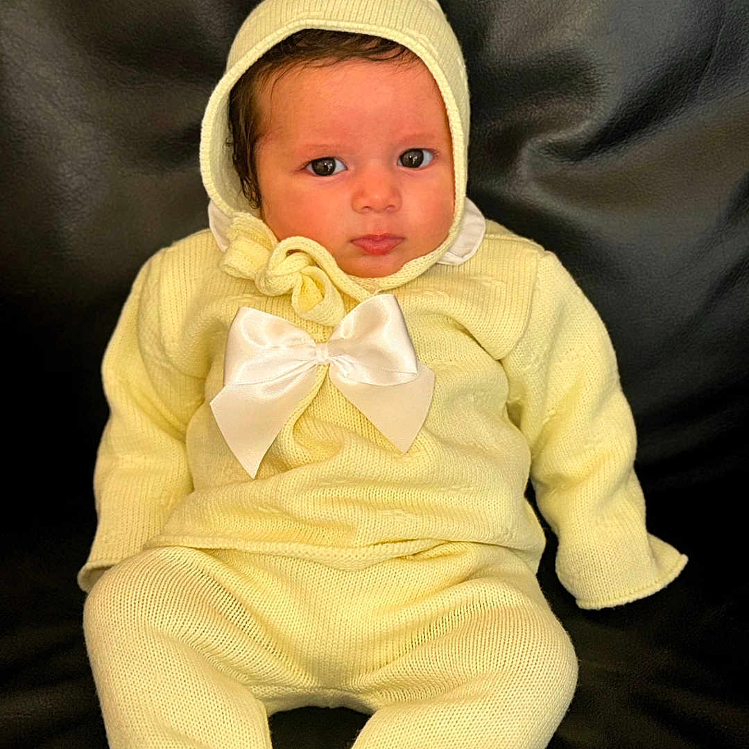 Rachel a rejoint le concours — aidez-le/la à gagner de superbes lots ! baby, infant, yellow_outfit, knitwear, bow, hood, sitting, black_leather, couch, cute, serious, portrait, child, indoors, warm_clothing, small_person, cozy, young, face, person