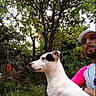 dog, man, outdoor, garden, tree, greenery, animal, pet, portrait, white_dog, black_spot, cap, casual_clothing, nature, foliage, summer, side_view, human, companion, leisure