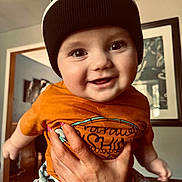 Alex joined the competition — help win amazing prizes! baby, child, smiling, face, hand, beanie, hat, indoor, person, clothing, orange_shirt, patterned_shorts, happy, cute, portrait, young_child, human, holding, home, living_room