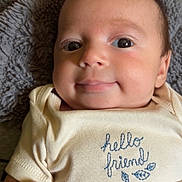 Clay is registered to the contest to win money with this photo: baby, infant, face, eyes, smile, hand, blanket, onesie, clothing, embroidery, text_on_clothing, skin, head, person, portrait, indoor, cute, young_child, human, cozy