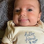 baby, infant, face, eyes, smile, hand, blanket, onesie, clothing, embroidery, text_on_clothing, skin, head, person, portrait, indoor, cute, young_child, human, cozy