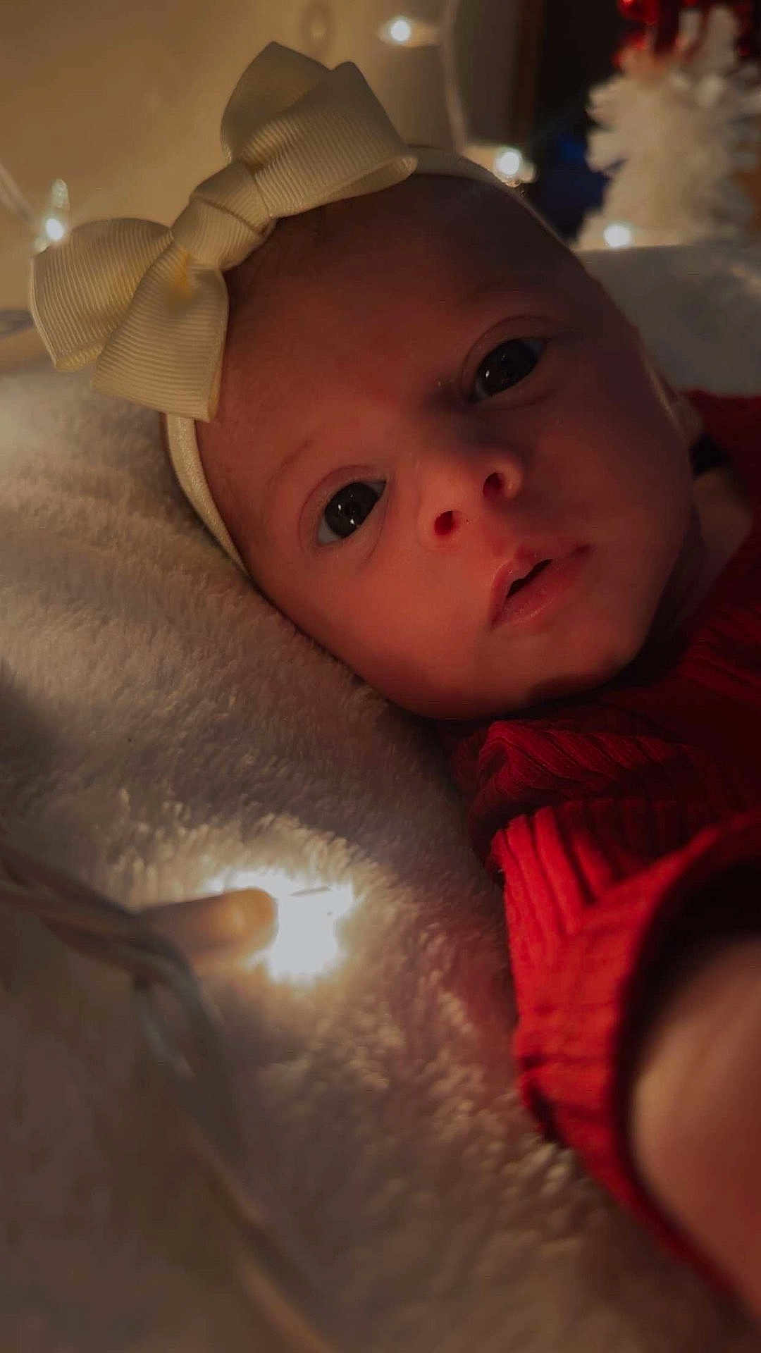 Raven joined the competition — help win amazing prizes! baby, infant, face, headband, bow, red_clothing, blanket, fairy_lights, soft_texture, closeup, portrait, indoors, cute, child, young, sleeping_area, warm_lighting, expression, cozy, holiday