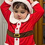 child, santa_costume, red_clothing, hat, indoor, curly_hair, face_paint, holiday, festive, buttoned_coat, white_fur_trim, belt, wooden_door, radiator, cute, portrait, young_child, smile, hands_up, costume