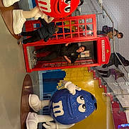 Zoe participe au concours pour gagner de l'argent avec cette photo : m&m, red_m&m, blue_m&m, child, telephone_booth, indoor, people, stairs, glass_railings, colorful_mural, floor, clothing, footwear, curiosity, display, shopping_area, casual, person, statue, toy