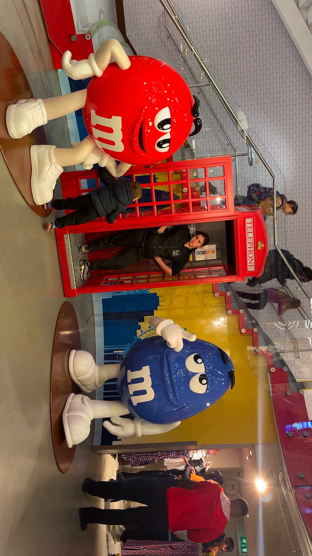 Zoe participe au concours pour gagner de l'argent avec cette photo : m&m, red_m&m, blue_m&m, child, telephone_booth, indoor, people, stairs, glass_railings, colorful_mural, floor, clothing, footwear, curiosity, display, shopping_area, casual, person, statue, toy