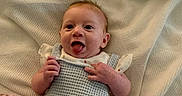 Hazel joined the competition — help win amazing prizes! baby, infant, blanket, knitted_clothing, smiling, tongue_out, lying_down, cute, happy, cozy, fabric, soft_texture, child, person, newborn, playful, indoors, warm, portrait, resting