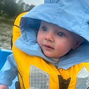 Teddy joined the competition — help win amazing prizes! baby, child, life_jacket, blue_hoodie, outdoor, water, boat, raincoat, hood, yellow, reflection, nature, person, portrait, young_child, curious, clothing, safety, closeup, face