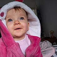 Adèle a rejoint le concours — aidez-le/la à gagner de superbes lots ! baby, child, hood, robe, pink, white, blue_eyes, smile, indoor, bed, blanket, curly_hair, person, infant, cute, soft_texture, cozy, warm_clothing, portrait, happy