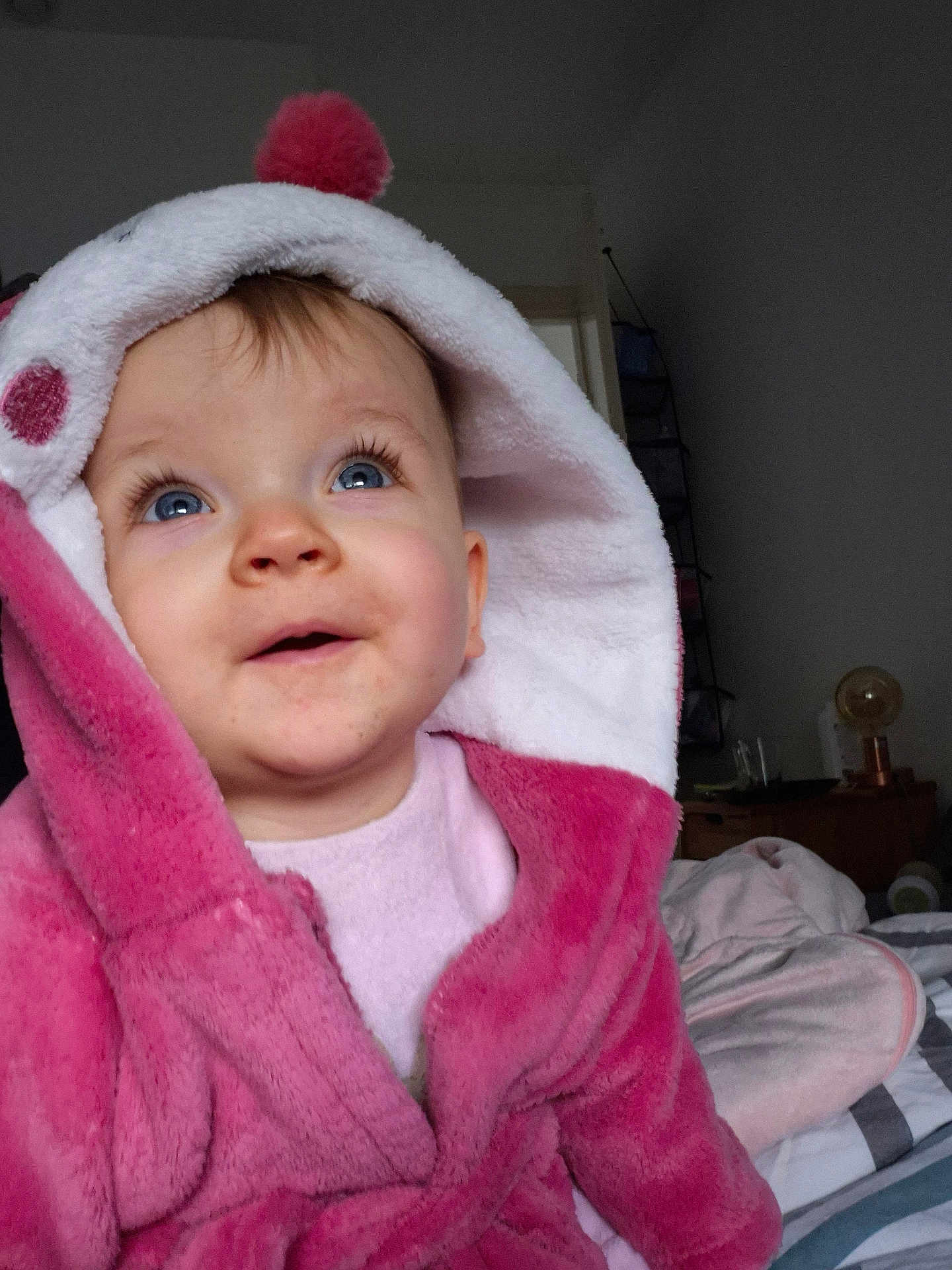 Adèle a rejoint le concours — aidez-le/la à gagner de superbes lots ! baby, child, hood, robe, pink, white, blue_eyes, smile, indoor, bed, blanket, curly_hair, person, infant, cute, soft_texture, cozy, warm_clothing, portrait, happy