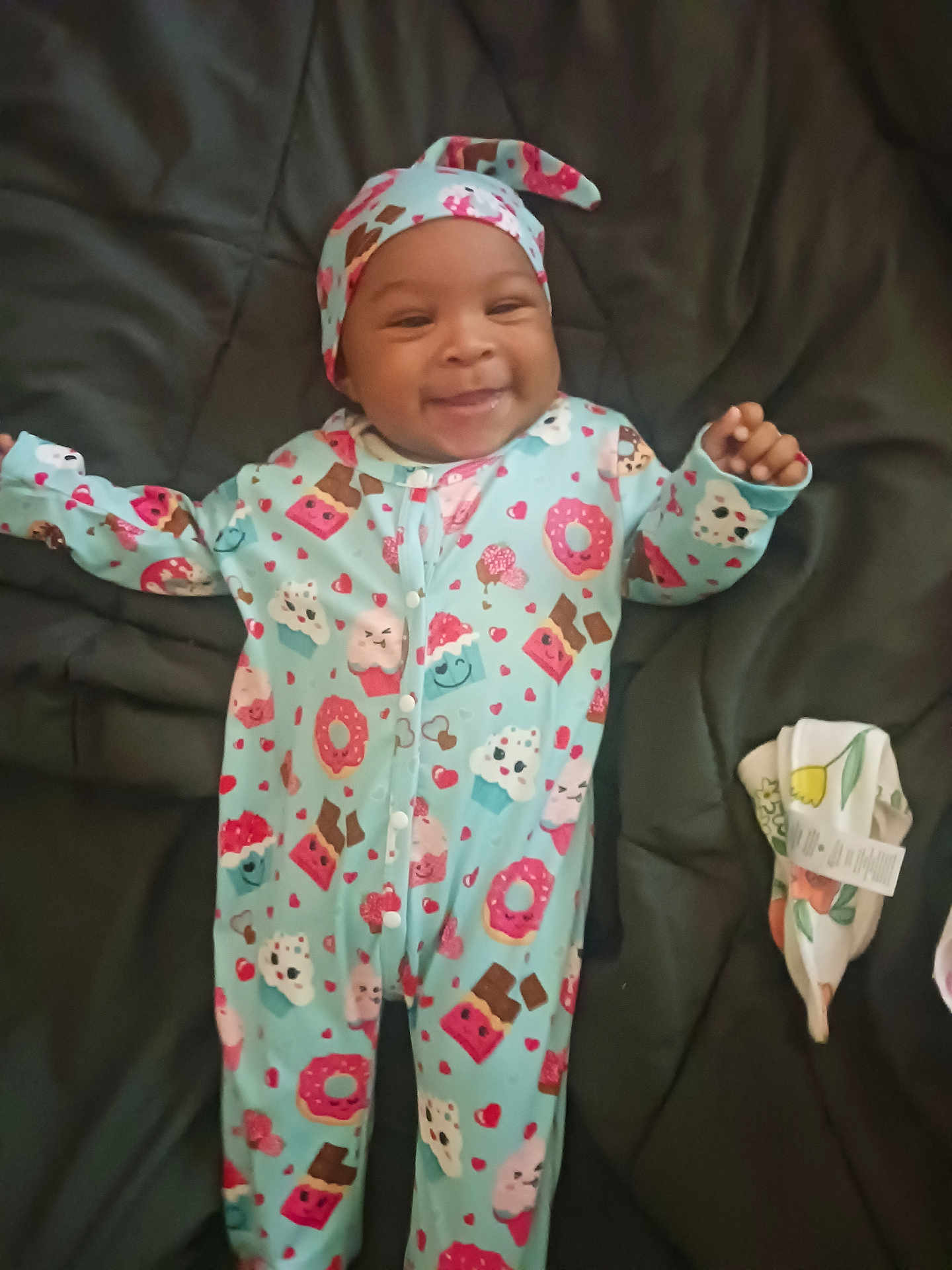 Kymani is registered to the contest to win money with this photo: baby, infant, smiling, onesie, hat, cute, lying_down, patterned_clothing, donuts, cupcakes, hearts, dark_background, comfort, happy, child, fabric, floral, soft, cozy, playful