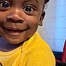 child, toddler, smiling, yellow_shirt, face, indoors, happy, person, closeup, cute, bright_color, eyes, expression, wall, clothing, portrait, baby, skin, hand, room